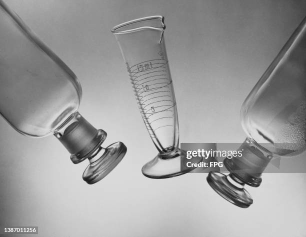 Close-up of a graduated measuring laboratory beaker between two bottles, each with glass stoppers, against a neutral background, circa 1955.