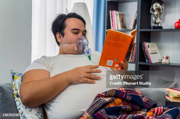 un jeune homme est assis dans un canapé avec un masque à oxygène et un livre de lecture - équipement d'assistance respiratoire photos et images de collection
