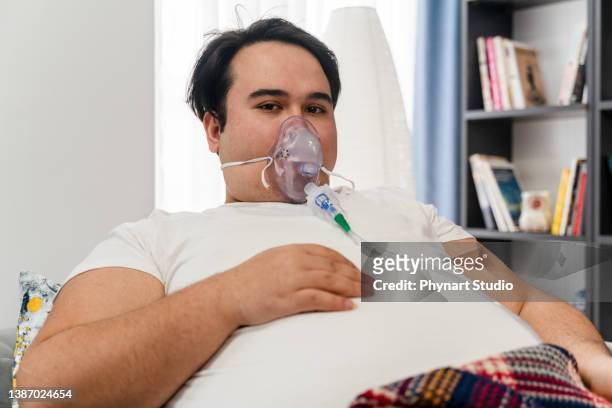 un jeune homme est assis dans un canapé avec un masque à oxygène et regarde la caméra - équipement d'assistance respiratoire photos et images de collection