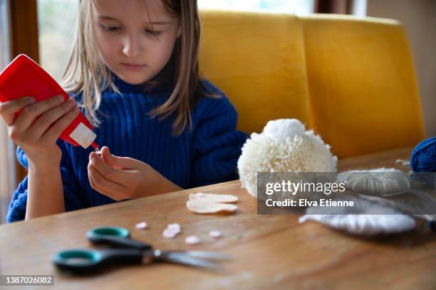 child (8-9) squeezing glue out of a plastic container and gluing pieces of felt onto homemade pom-pom crafts from cream coloured wool. - sticky stock pictures, royalty-free photos & images