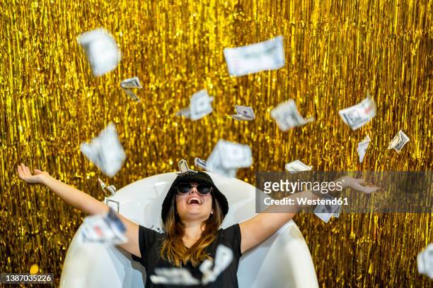 carefree woman lying in bathtub with arms outstretched amidst falling money - geld stock-fotos und bilder