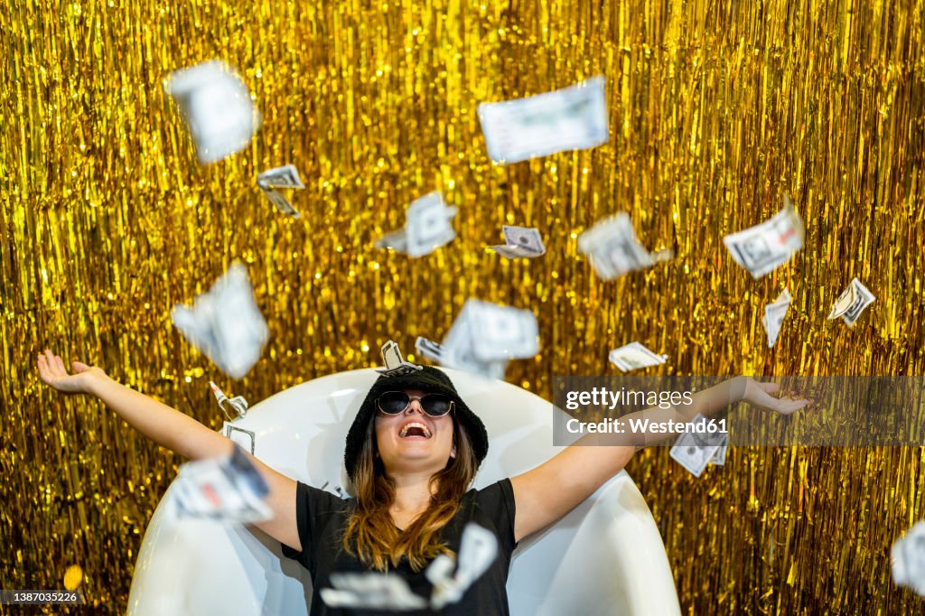 Carefree woman lying in bathtub with arms outstretched amidst falling money