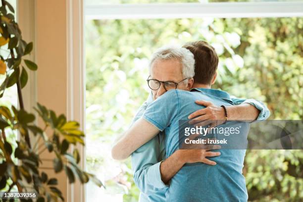 senior man hugging son against window at home - mid adult men stock pictures, royalty-free photos & images
