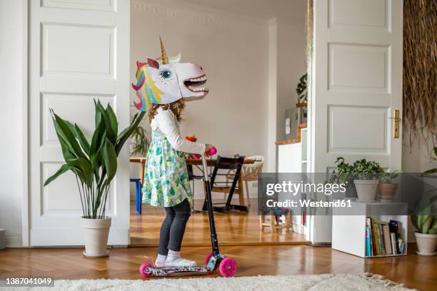 girl with unicorn mask riding push scooter at home - tretroller stock-fotos und bilder