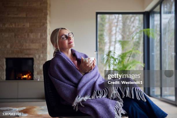 relaxed woman wrapped in blanket sitting with eyes closed in living room - wrapped in a blanket stock pictures, royalty-free photos & images