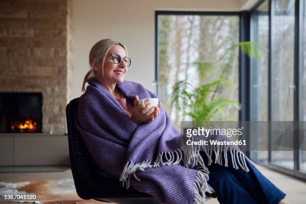 happy blond woman wrapped in blanket holding coffee cup sitting at home - blanket stock pictures, royalty-free photos & images
