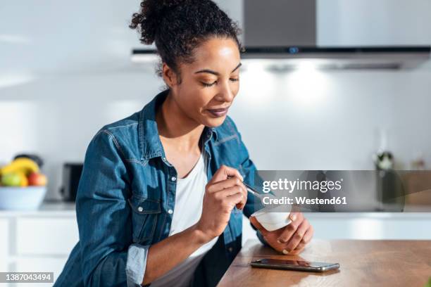 woman eating yogurt sitting in kitchen at home - yogurt foto e immagini stock