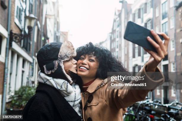 happy young woman taking selfie with friend kissing on cheek in city - friends kissing cheeks stockfoto's en -beelden