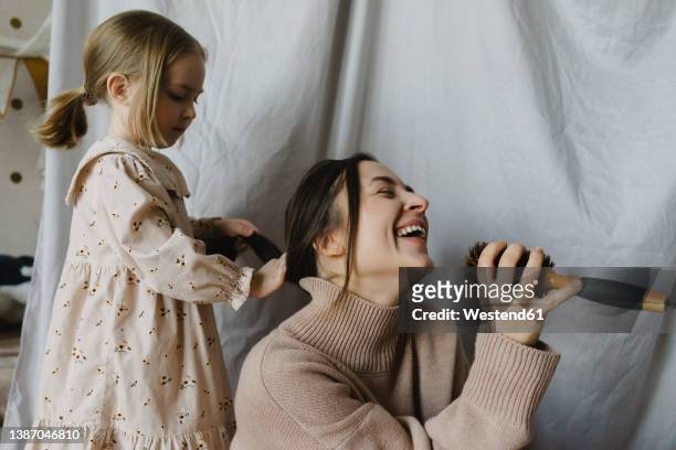 cheerful woman holding hairbrush singing by daughter tying hair at home - combing ストックフォトと画像