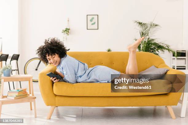 happy young woman surfing net through smart phone lying on sofa in living room at home - ligga på mage bildbanksfoton och bilder