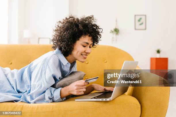 smiling woman holding credit card using laptop lying on sofa in living room at home - online shopping foto e immagini stock