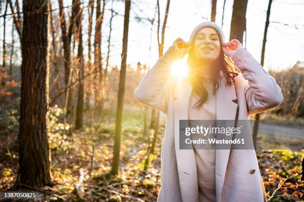 happy woman in knit hat standing by trees in forest on sunny day - winter coat stock pictures, royalty-free photos & images