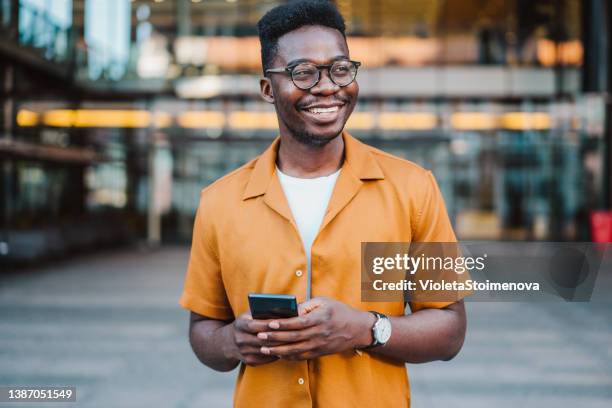 joven sonriente usando teléfono inteligente en la calle. - casual man walking fotografías e imágenes de stock