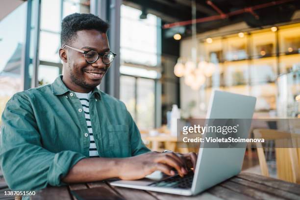young man sitting in coffee shop and using laptop. - african creative with laptop working outside stockfoto's en -beelden