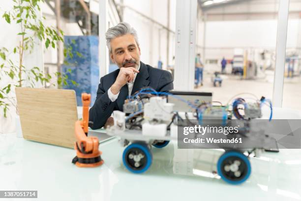 businessman sitting with hand on chin looking at model of robotic vehicle in factory - prototype stock pictures, royalty-free photos & images