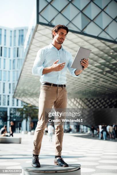 smiling businessman using tablet pc standing on seat outside modern building - mann vor pc stock-fotos und bilder