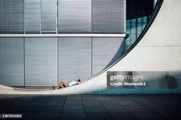 businessman with hand behind head lying on wall in front of modern building - the berlin wall stock-fotos und bilder