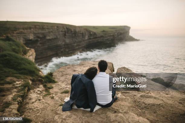 newlywed couple sitting by the sea at sunset - styles stock pictures, royalty-free photos & images