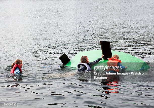 three children swimming around capsized sailing dinghy - capovolgersi foto e immagini stock