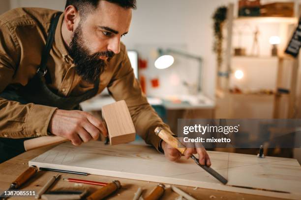 bearded artist carving wood with chisel and hammer in studio - fries stockfoto's en -beelden