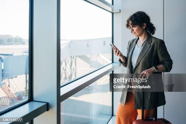 a smiling caucasian female standing at the airport and holding her luggage while watching something on her smartphone - passenger stock pictures, royalty-free photos & images