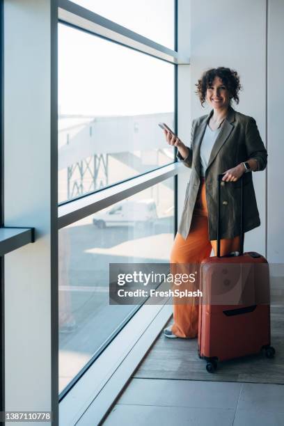 a smiling caucasian female standing at the airport and holding her luggage in one and a smartphone in the other hand - carry on bag imagens e fotografias de stock