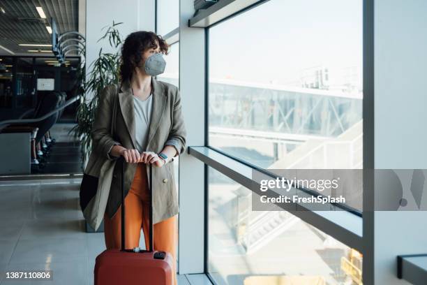 travelling during pandemic: a pensive businesswoman wearing a mask against coronavirus holding her luggage while waiting at the airport to board a plane - airport mask stock pictures, royalty-free photos & images