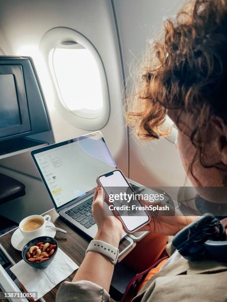 eine unkenntliche kaukasische frau, die im flugzeug sitzt und ihr smartphone benutzt, während ihr laptop im hintergrund eingeschaltet ist - flying reading stock-fotos und bilder