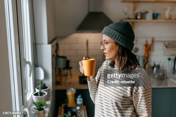 woman with coffee cup looking through window at home - woman drinking tea stockfoto's en -beelden