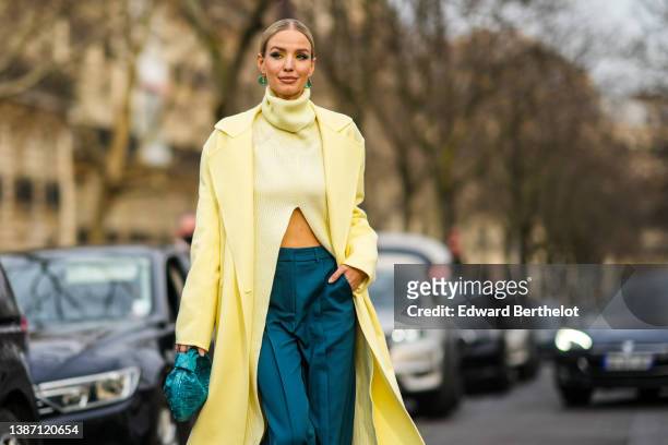 Leonie Hanne wears gold and green pendant earrings, a pale yellow ribbed turtleneck / asymmetric cropped pullover, a pale yellow long coat, high...