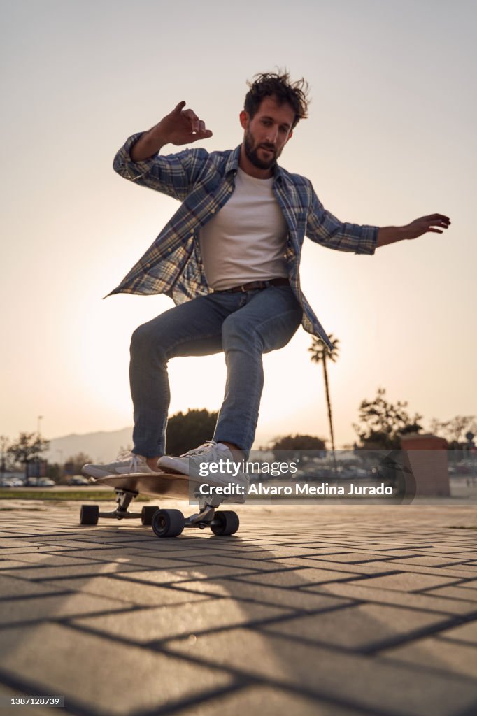View from below of a young ombre dressed in casual clothes skateboarding on a longboard in the city at sunset.
