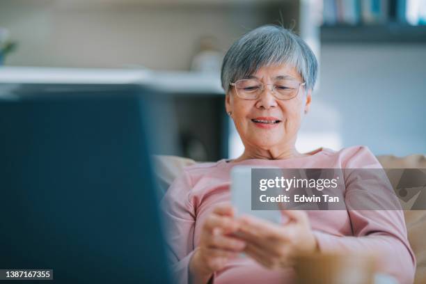 asian chinese senior woman using smart phone reading text message in living room of her apartment in city - asian old woman stock pictures, royalty-free photos & images