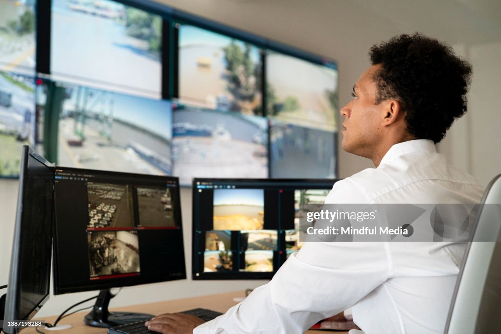 Young adult male security worker watching video wall while sitting at desk