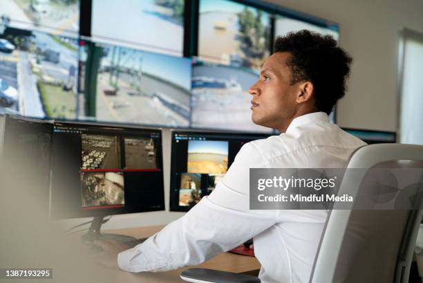 jovem adulto homem trabalhador de segurança assistindo parede de vídeo enquanto sentado na mesa - sala de controle - fotografias e filmes do acervo