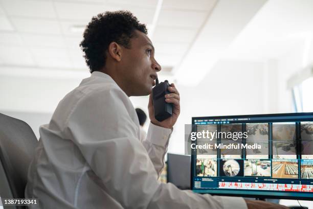 young adult male security worker using walkie talkie while looking at security camera images on computer monitor - guarding stock pictures, royalty-free photos & images