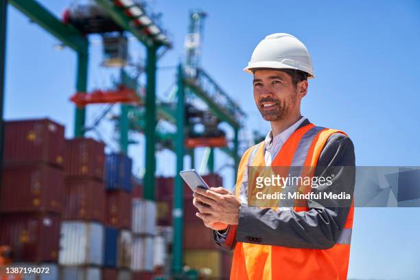 asian american male engineer using smart phone while standing at shipping container yard - funcionário de alfândega imagens e fotografias de stock
