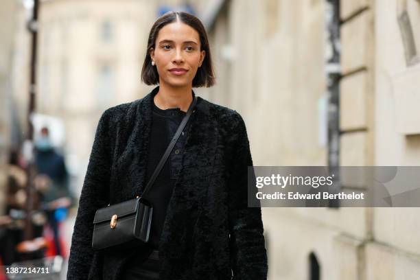 TyLynn Nguyen wears a black ribbed polo shirt, a black fur long coat, a black shiny leather crossbody bag, a black shiny leather belt, black suit...