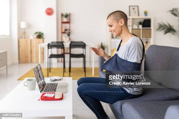 woman in an arm sling using a laptop while having a video call with her doctor at home. - at home health care stock pictures, royalty-free photos & images