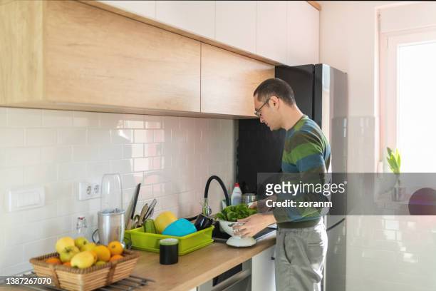 young man standing by the kitchen sink at home with strainer full of lettuce - colander stockfoto's en -beelden