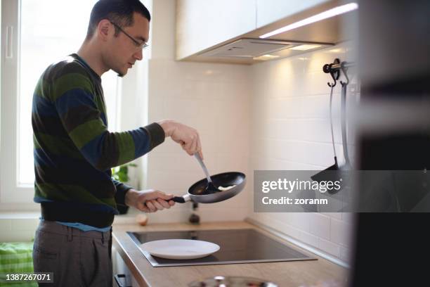 ヘラの入った鍋から目玉焼きを拾い上げて皿に盛り付ける青年 - electric stove burner ストックフォトと画像