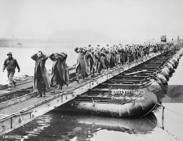 With their hands above their heads, prisoners of war from units of the German 11th SS Panzer Army walk into captivity along a pontoon bridge across...