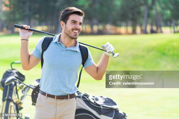 smiling young golf player holding golf club on his shoulders, standing next to his clubs in a bag on a golf course. - billionaire stock pictures, royalty-free photos & images