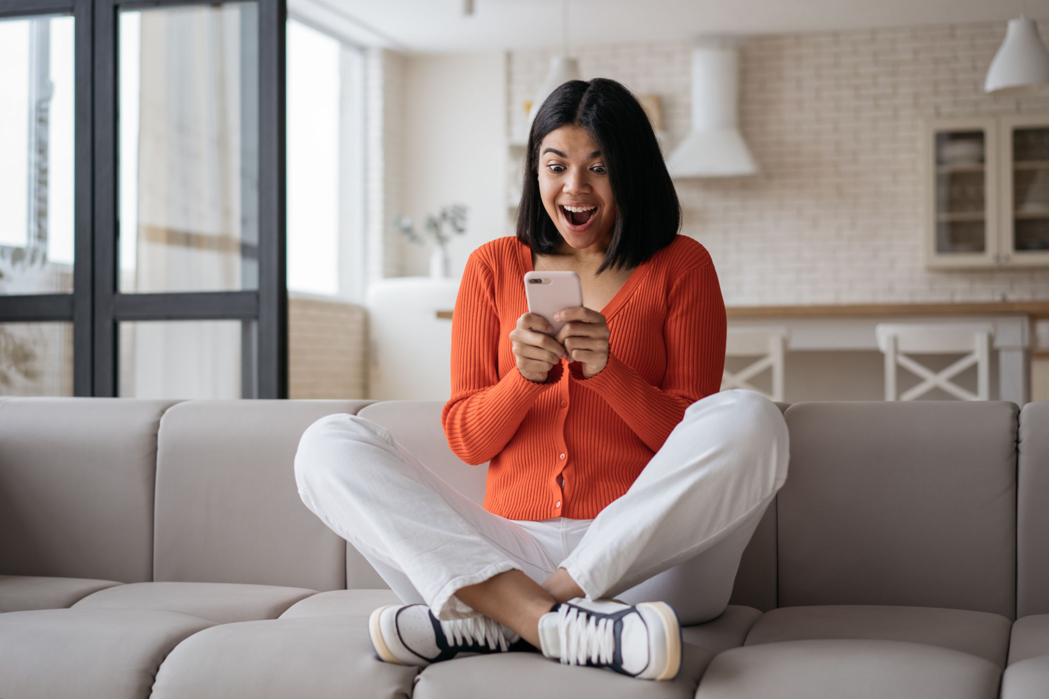 Woman holding smartphone playing mobile game celebration success at home. Excited African American hipster using mobile phone shopping online with big sales sitting on sofa Woman holding smartphone playing mobile game celebration success at home. Excited African American hipster using mobile phone shopping online with big sales sitting on sofa
