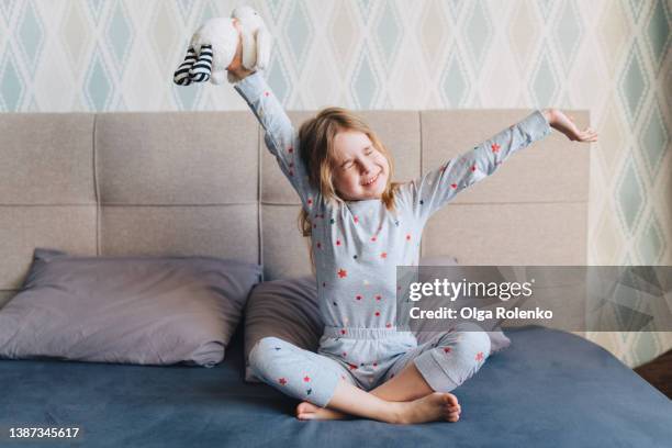 funny and pretty little blond, long-haired girl awakening and sitting on a comfortable bed with her favorite stuffed toy rabbit. - schneidersitz stock-fotos und bilder