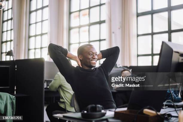 smiling male computer programmer sitting with hands behind head at desk in office - hands behind head stock pictures, royalty-free photos & images
