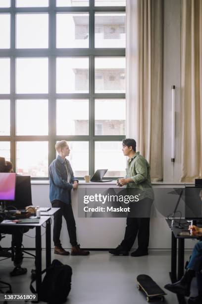 side view of male computer programmers talking while standing by window in office - coffee break office ストックフォトと画像