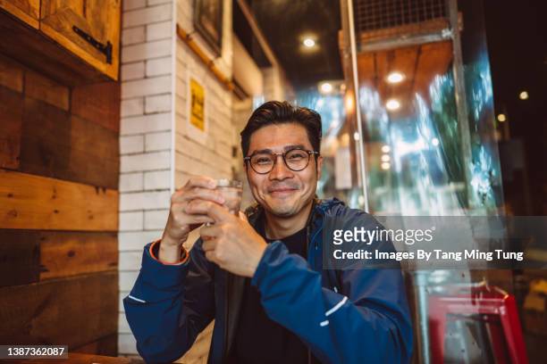 young handsome asian man holding a glass of water, smiling joyfully at the camera in a restaurant - sitting at table looking at camera stock-fotos und bilder
