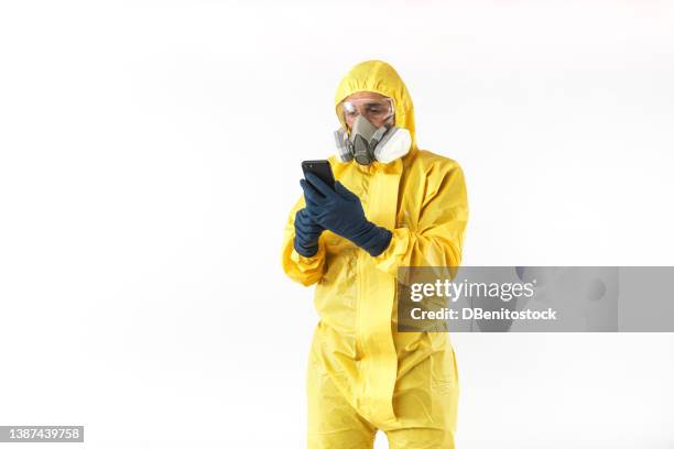 technician with yellow nuclear protection suit, mask and protective glasses consulting his mobile phone, on white background. concept of nuclear energy and pandemics. - schutzanzug stock-fotos und bilder