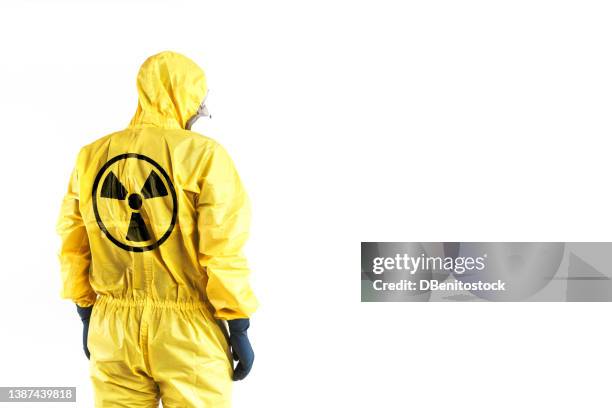 technician in a yellow nuclear protection suit, mask and protective glasses, with his back to the nuclear symbol, on a white background. concept of nuclear energy and pandemics. - radioactive warning symbol photos et images de collection