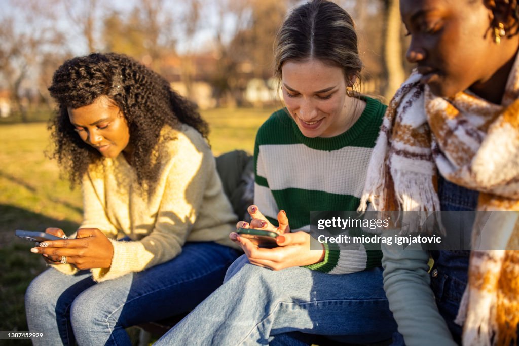 Modern multiracial female friends, sitting on the park bench and surfing the net on mobile phone